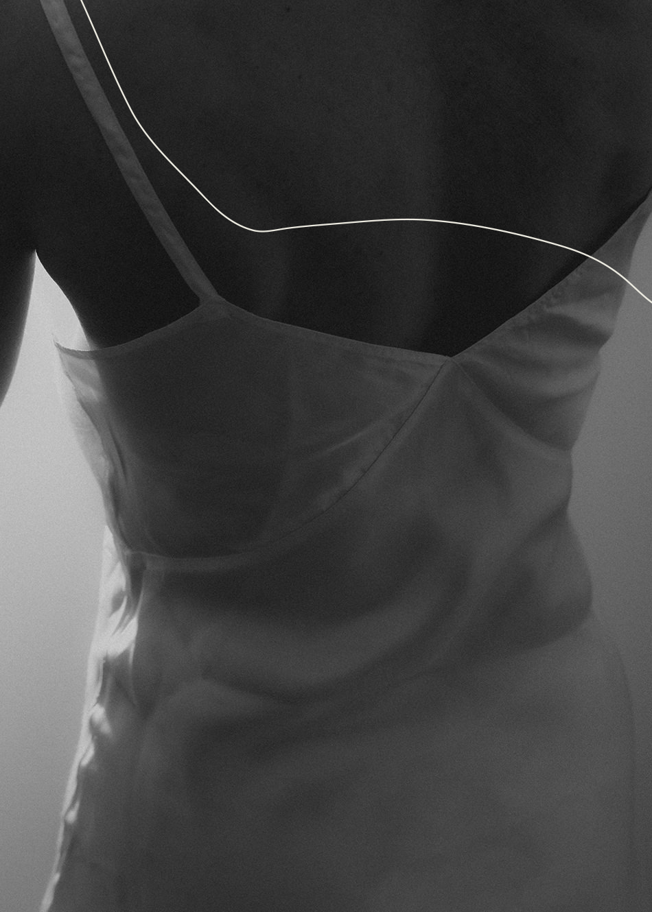 A grayscale photo shows the back of a person wearing a satin slip dress with thin straps, highlighting the fabric’s smooth texture and soft lighting. A thin, curved white line is superimposed across the upper part of the image.