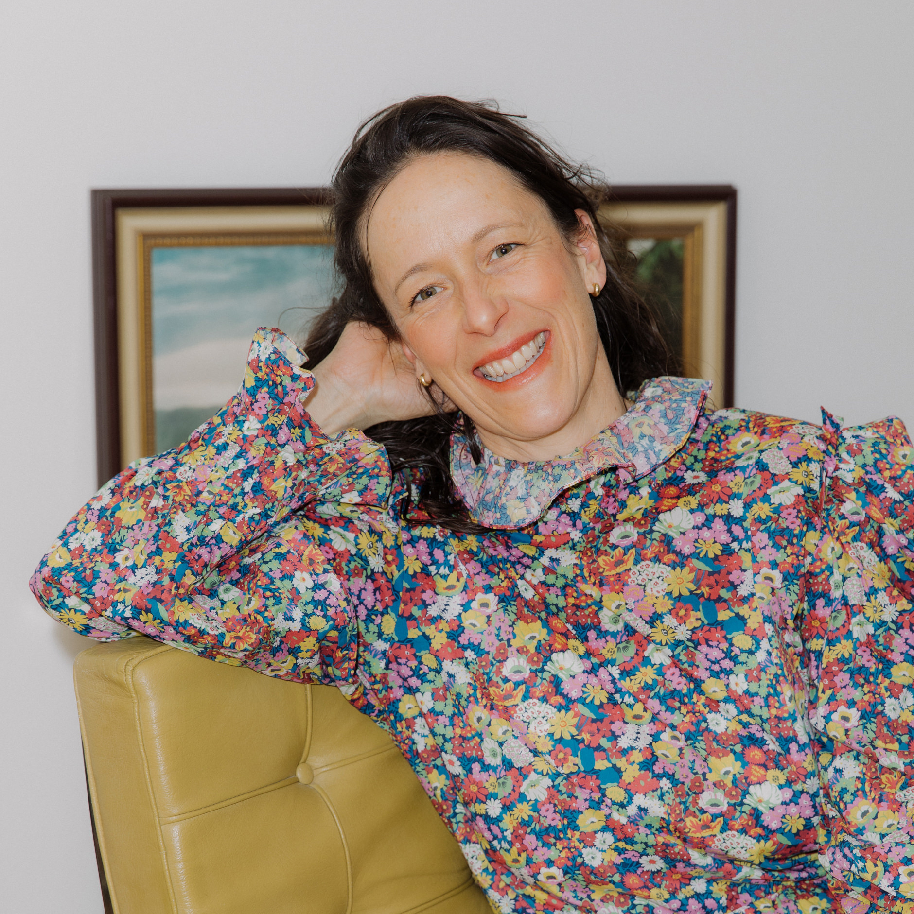 A woman with long dark hair, wearing a colorful floral dress, smiles while sitting on a yellow chair. A framed painting is visible on the white wall behind her.